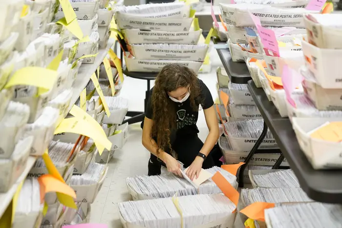 A worker sits in the middle of dozens of bins of absentee ballots